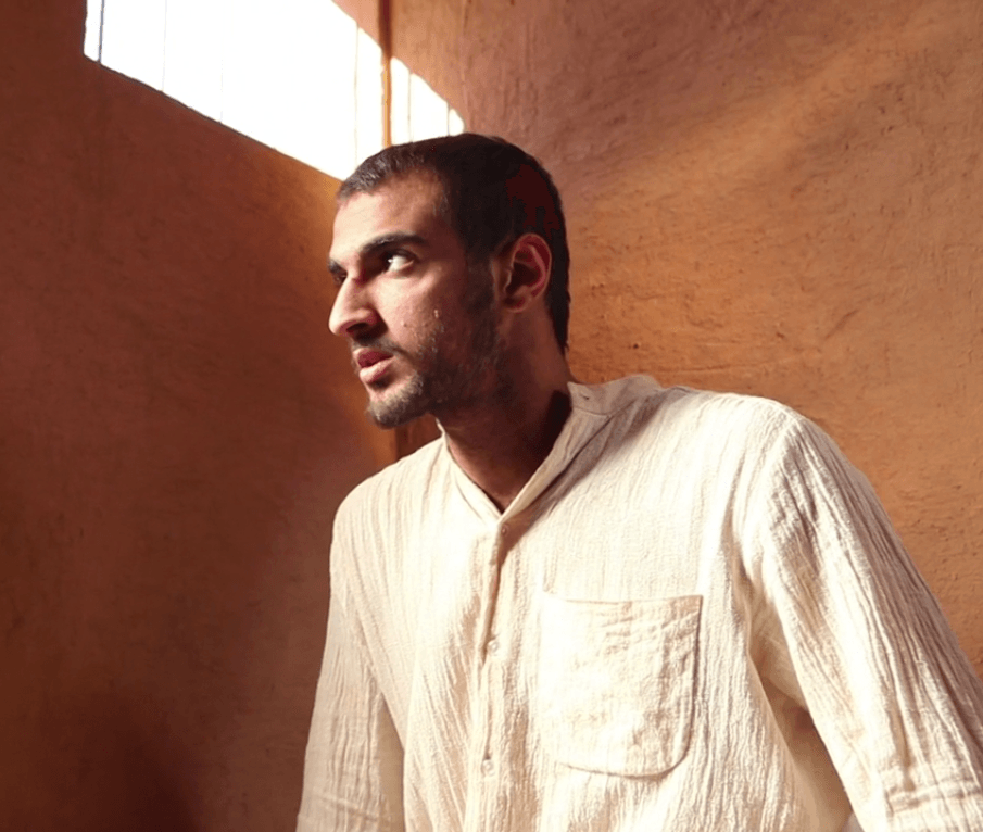 A person in a light-colored shirt stands in a sunlit, textured room. They are looking thoughtfully towards the left. The walls have a warm, earthy tone with light filtering in from a window above.